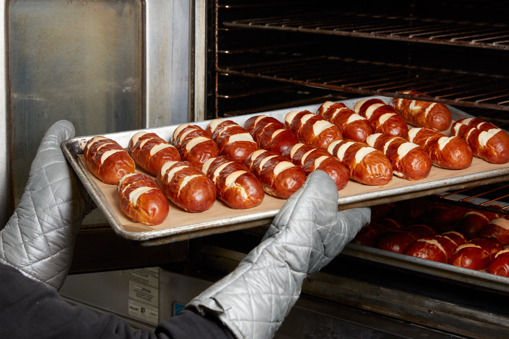 A baker in oven mitts pulling a sheet tray of golden-brown pretzel rolls from a commercial oven