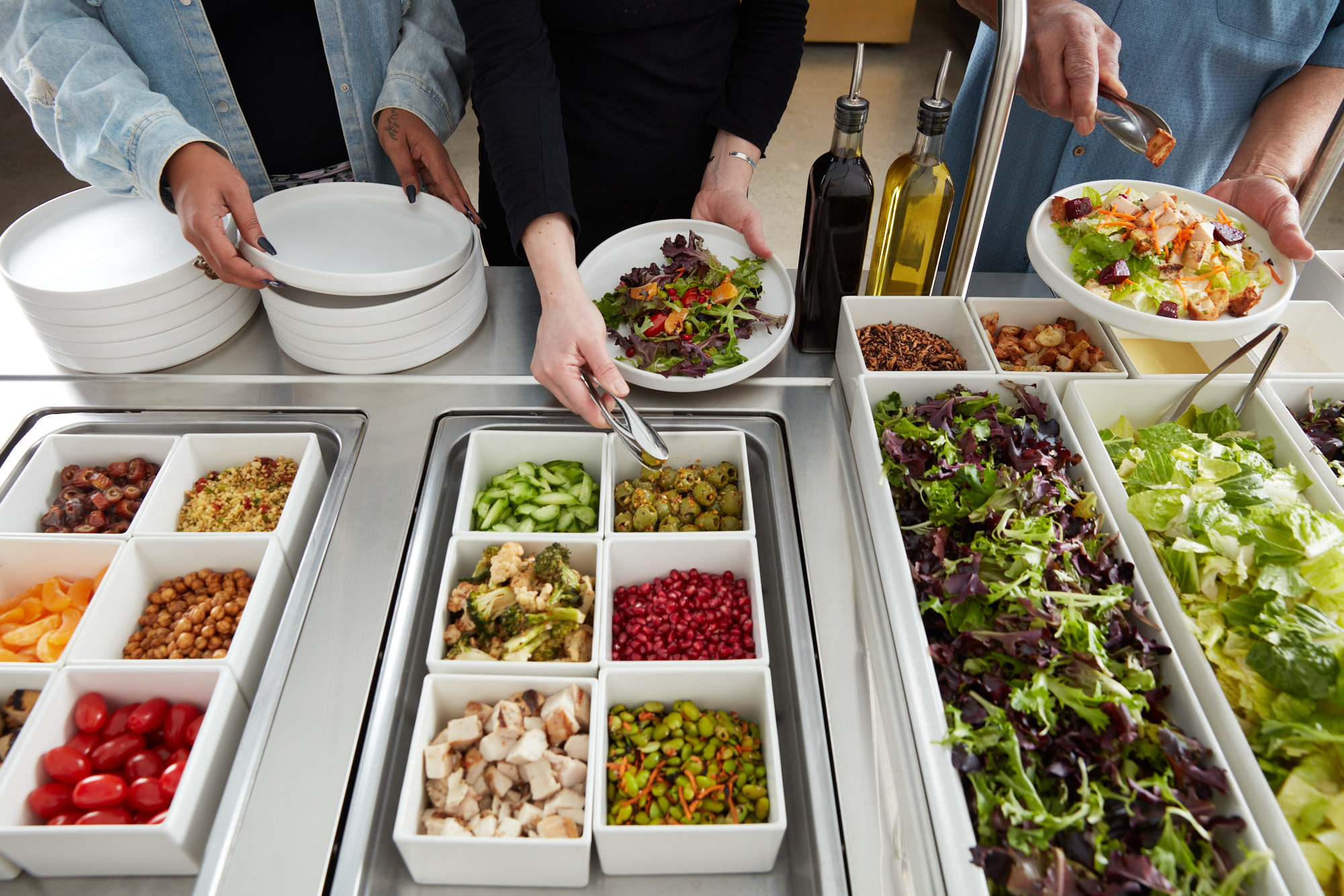 An overhead build-your-own salad bar with white bins of fresh toppings including tomatoes, chickpeas, cucumbers, olives, pomegranate, and greens